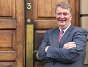 Man in suit stands in front of wooden doors with arms folded and smiling
