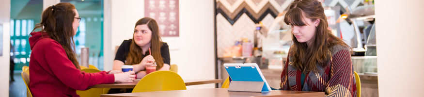 3 students in cafe, two students talk to each other one reading something on tablet