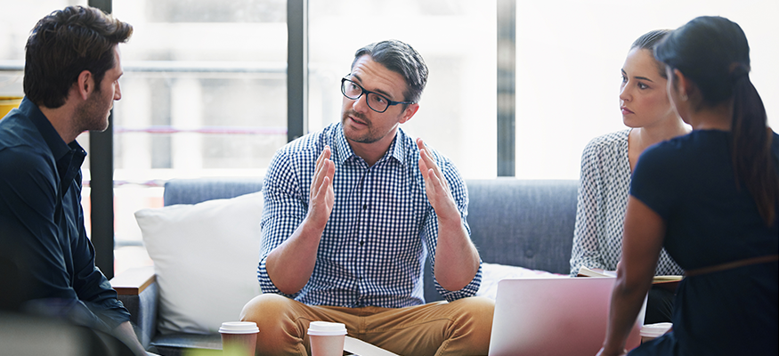 A man sat on couch addressing a group of employees sat around him