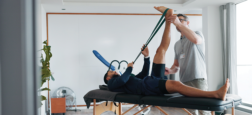 A man lying on a treatment table, supported by a male Physiotherapist as he uses resistance bands to stretch his leg into the air.