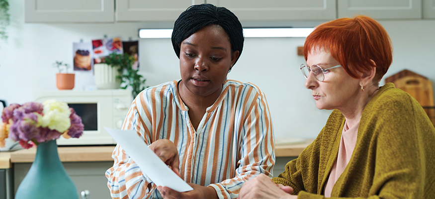 A social worker helps a woman by reviewing a document with her at her kitchen table.
